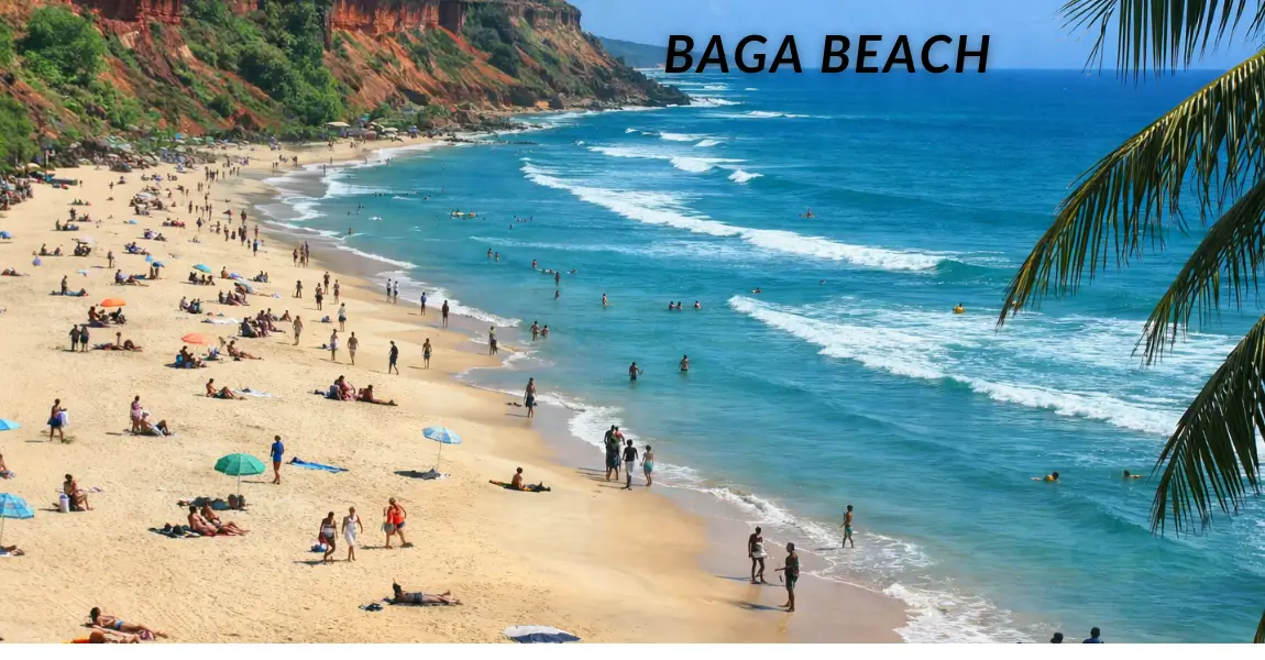 Wide-angle view of a crowded Goa beach with golden sand, turquoise waves, red cliffs and palm trees under clear blue sky